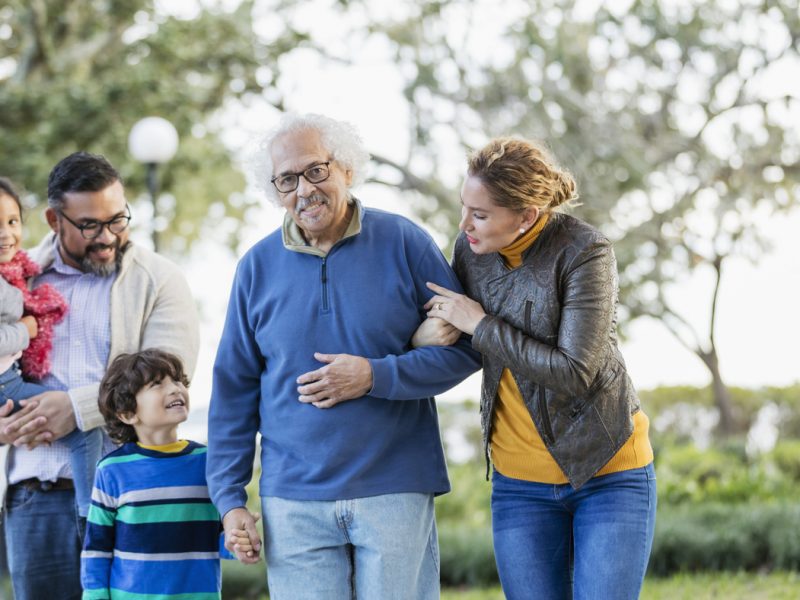 A multi-generation Hispanic family standing together outdoors in a park. The grandfather is in his 80s, standing arm in arm beside his adult daughter, in her 30s, and holding his grandson's hand. The boy is 6 years old and his sister is 4.